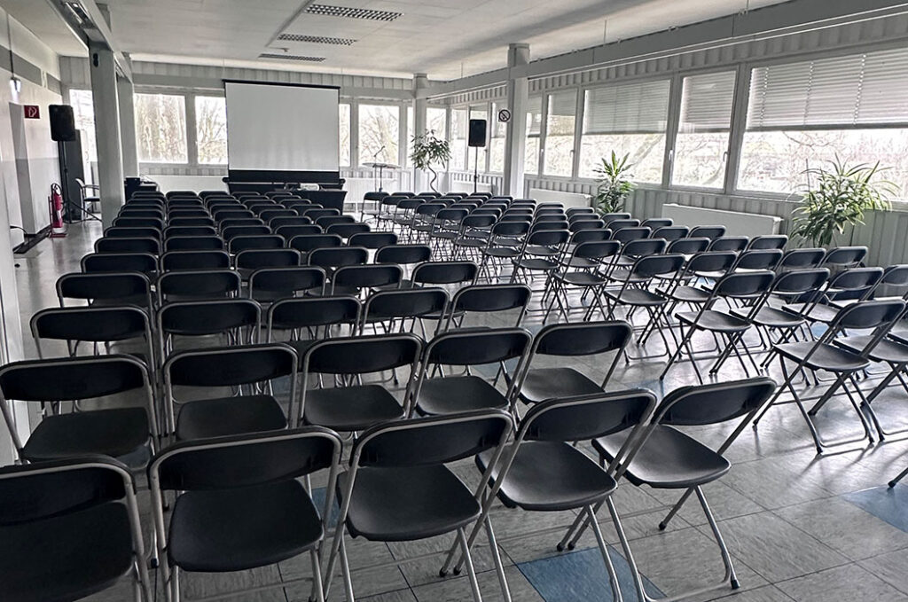 In einem hellen Raum stehen viele schwarze Klappstühle vor einer weißen Leinwand