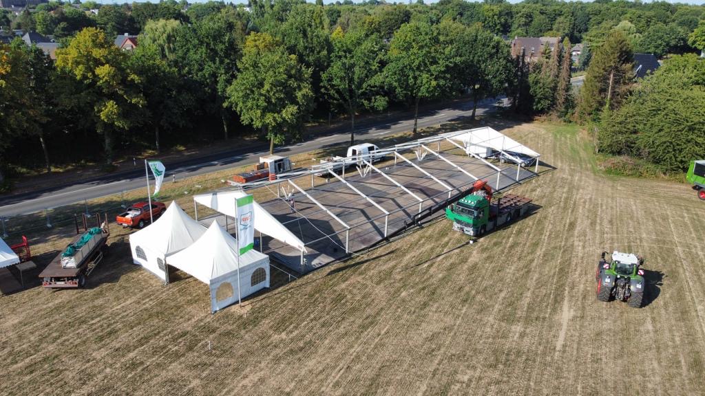 In einem großen Zeltaufbau auf braunem Feld stehen mehrere Fahrzeuge und Arbeiter mit neutralem Gesichtsausdruck