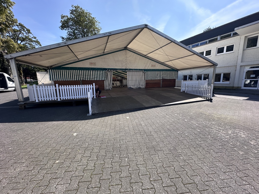 Ein großes Zelt mit weißem Dach steht auf einem gepflasterten Platz vor einem Gebäude
